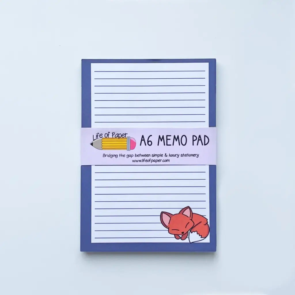 An Fox Memo Pad with a blue border and a sleeping red fox illustration in the bottom right corner. The pad, which is fountain pen friendly, is wrapped with a band featuring a pencil design and the text "Life of Paper, A6 Memo Pad, Bridging the gap between simple & luxury stationery www.lifeofpaper.com.