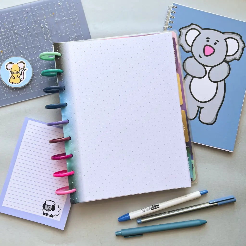 A workspace featuring a dotted notebook with colorful tabs and rings, a blue notebook with a cartoon koala on the cover, a small notepad with a cartoon sheep, various pens in blue and white, and a clear pencil case with planner accessories including Galaxy Themed Notepaper and a sticker.