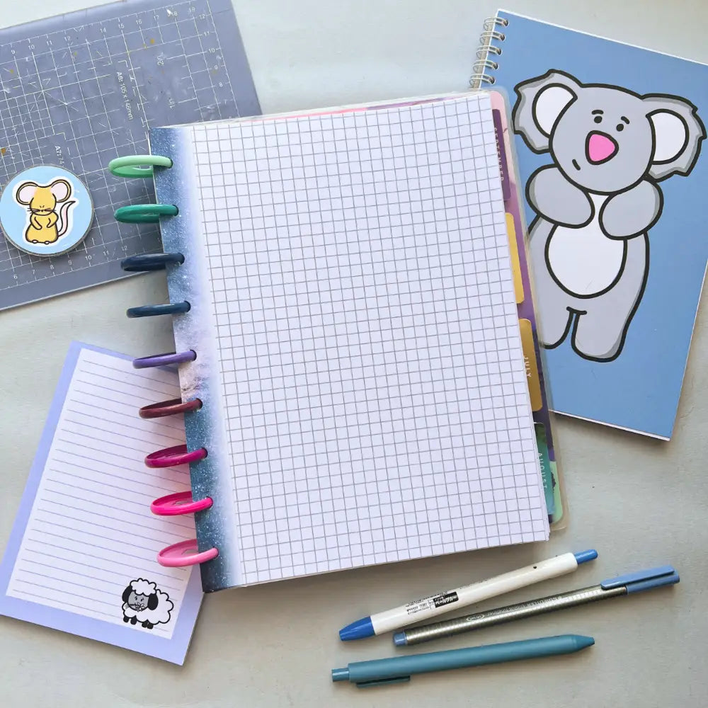An open graph paper notebook with colorful pens clipped on the side sits next to a blue notebook featuring a cartoon koala cover, a sticker of a mouse, and a small notebook with a sheep illustration. Three pens lie nearby on the light gray surface, all complemented by Galaxy Themed Notepaper.