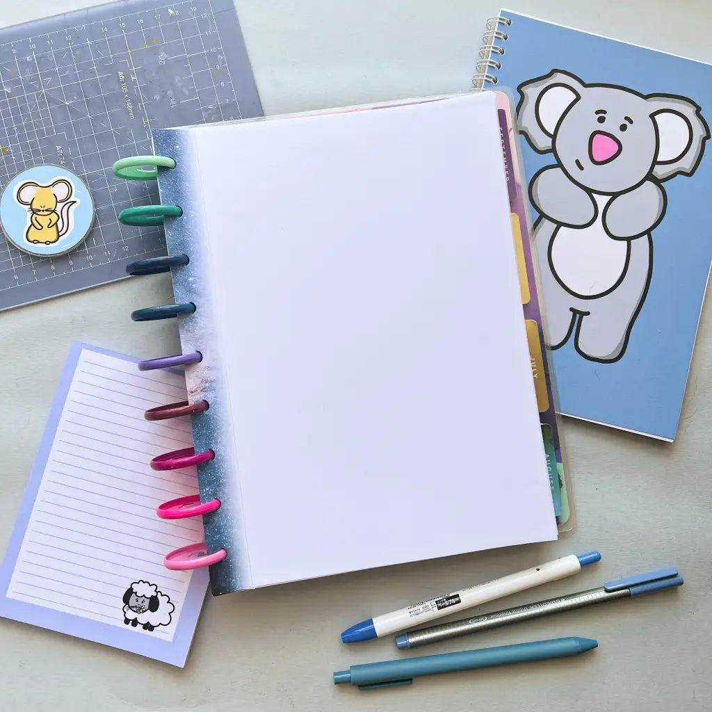 A flat lay of stationery items featuring a notebook with rainbow-colored rings, a blue notebook adorned with a cartoon koala, and Galaxy Themed Notepaper. Included are three pens and a plastic sheet with a mouse-in-a-cup sticker—ideal planner accessories for your galaxy star theme.