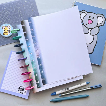 An open notebook with colorful tabs, a blue koala-covered notebook, a small purple note with a sheep illustration, two pens, and a Galaxy Themed Notepaper ruler with an animal sticker lie on the desk. The colorful tabs contrast with the white and blue paper, enhancing the workspace's vibrant feel.
