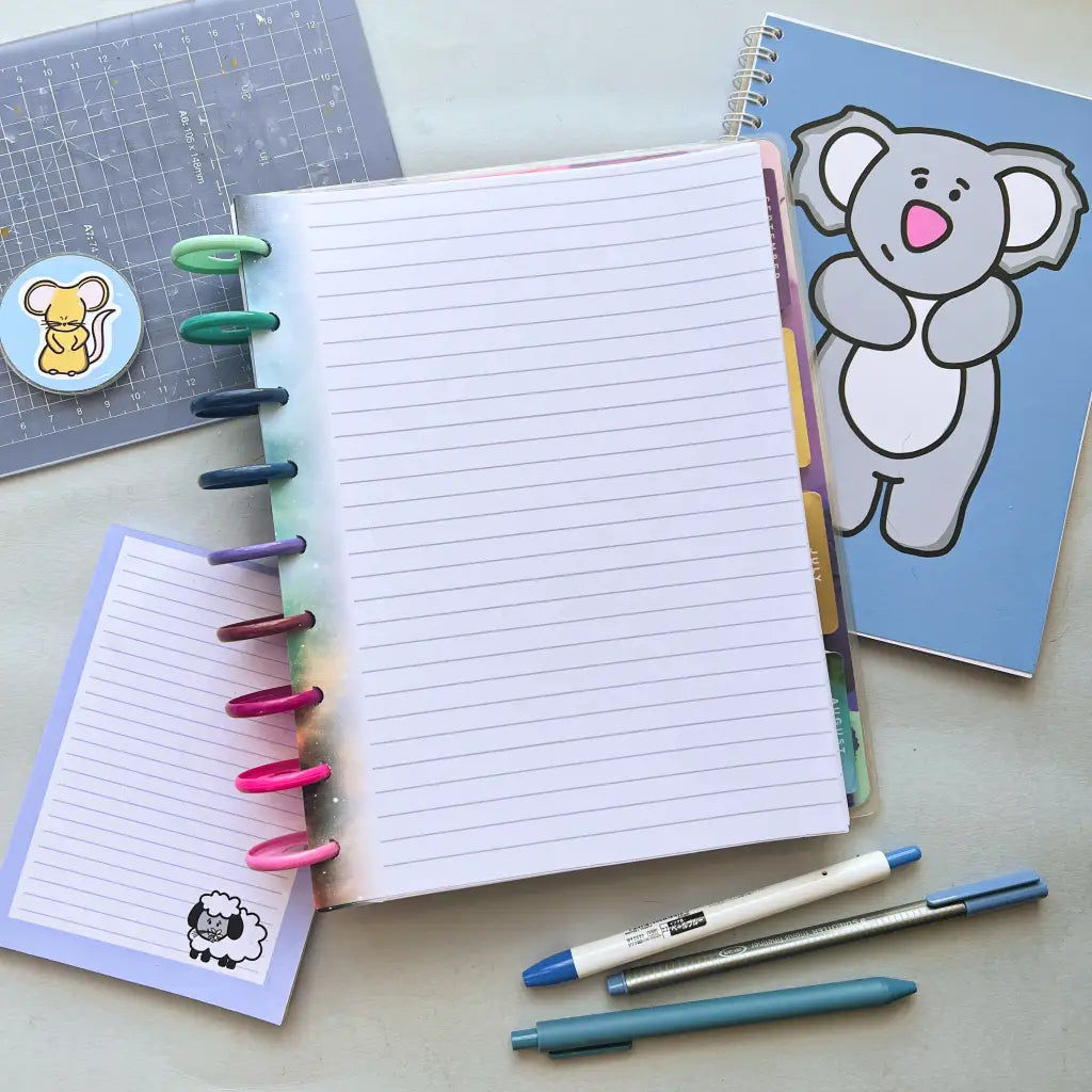 A colorful planner with rainbow rings lies open, showing Galaxy Themed Notepaper. Next to it, there's a blue notebook featuring a cartoon koala on the cover. A smaller lined notepad with a sheep sticker, stickers of a mouse, and a grid ruler are also on the table, along with three pens and other planner accessories.