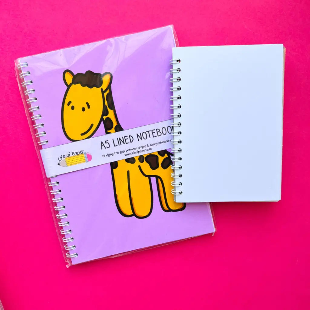 An A5 spiral-bound notebook with a cartoon giraffe cover lies on a bright pink surface next to a smaller plain white spiral-bound notebook. The Happy Giraffe Notebook features a cute illustration and a label that reads "A5 Lined Notebook, 40 pages.