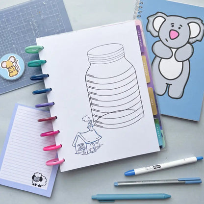 A desk setup features an illustrated notebook open to a page with a large jar drawing labeled "House Deposit Saving Tracker" and a small house at the bottom. Surrounding the notebook are color pens, a blue card with a koala design, another notepad with a sheep, and a cutting mat.