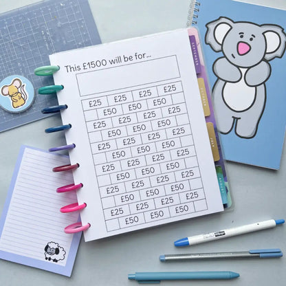 A finance planning notebook lays open with a budgeting page outlining allocations for £1500, featuring a personalised House Deposit Saving Tracker. Surrounding items include colorful pens clipped to the notebook spirals, a cartoon koala card, a pad with a sheep image, a blue pen, and a precision knife.