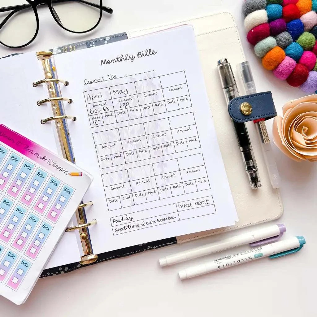 A neatly organized desk featuring a Household Bill Tracker in a ring binder. Nearby are pens, glasses, colorful stickers, a felt ball coaster, a peach-colored rose, and a pack of sticky notes. The Household Bill Tracker lists council taxes for April and May with columns for amounts and dates.