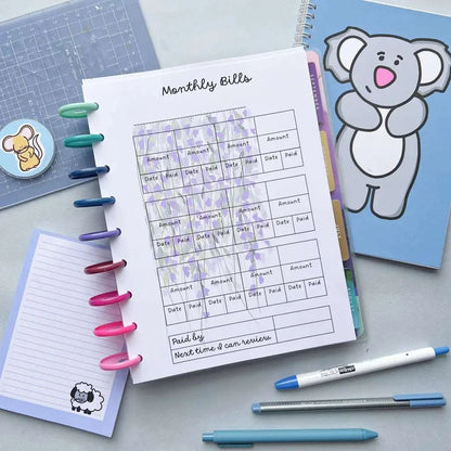 A "Household Bill Tracker" on a spiral notebook, surrounded by pens in various colors, a notebook with a koala on the cover, a small sheet of paper with a sheep illustration, and other stationery items on a gray surface.