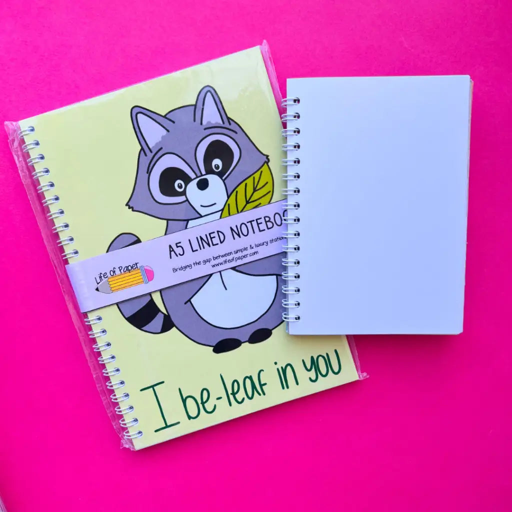A yellow I Be-leaf In You Notebook with an illustration of a raccoon holding a leaf and the text "I be-leaf in you" lies next to a plain white notebook. This encouraging notebook, along with its companion, features spiral bindings. The background is a bright pink color.