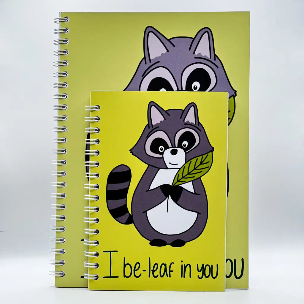 Two spiral raccoon notebooks stand side by side, each raccoon holding a green leaf. The smaller, I Be-leaf In You Notebook is in front, partially obscuring the larger one. These encouraging notebooks are perfect for brightening up your study sessions.