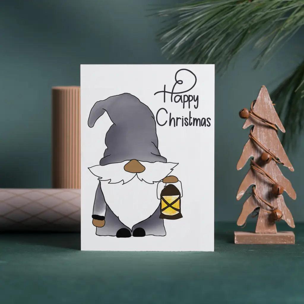 This delightful Gonk with Lantern Christmas Card showcases a Nordic Gonk sporting a grey hat and holding a lantern. "Happy Christmas" is elegantly scripted beside the gnome. The background includes festive decorations such as rolled paper and a wooden tree, all thoughtfully designed with biodegradable packaging.