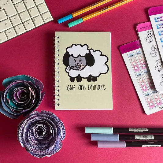 A flat lay photo of a desk with a keyboard, two colorful paper flowers, pens, and paper planners. A 40-page Mini Ewe Are Brilliant Notebook with a cute sheep illustration and the text "ewe are brilliant" is centered on a bright pink background.