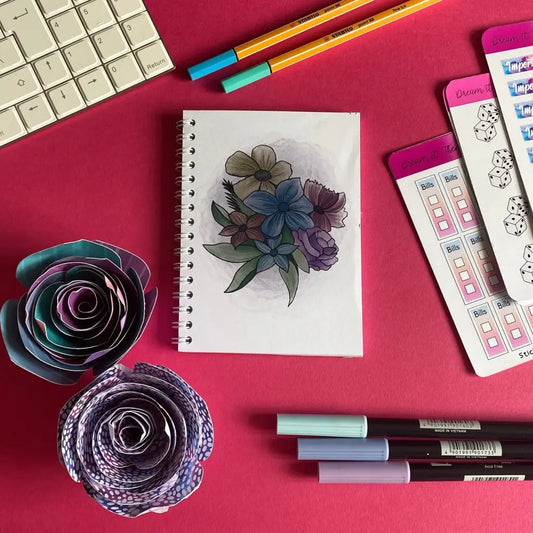 A pink workspace displays a keyboard, a Mini Faded Floral Notebook, handmade paper roses, pastel-colored markers, and a planner pad with checklists and dice illustrations. The colorful arrangement is neatly laid out on the desk.