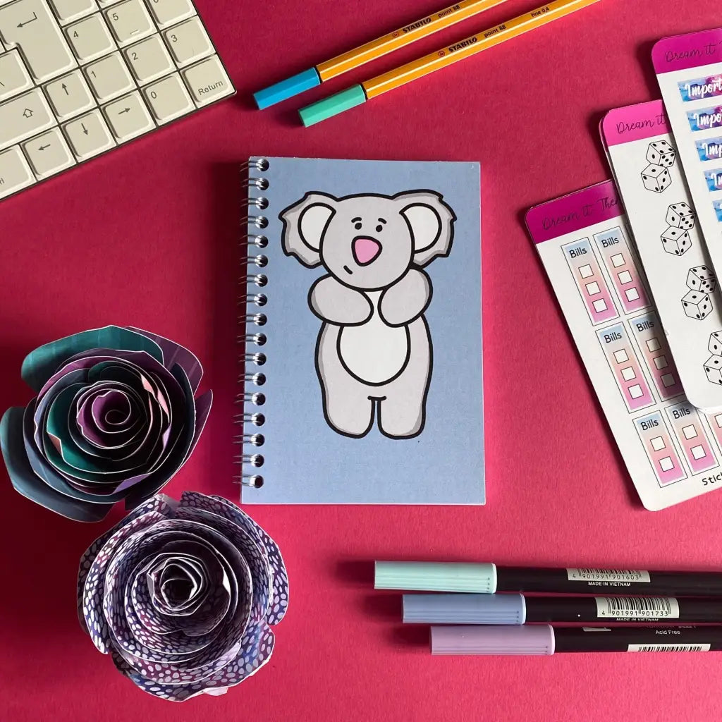 A Mini Gentle Koala Notebook with a cartoon koala on the cover sits on a pink surface. Around it are origami flowers, colorful pens, a partial keyboard, and stationery items, including stickers and a to-do list. Pencils and markers are scattered, adding a vibrant touch perfect for calm note-taking sessions.