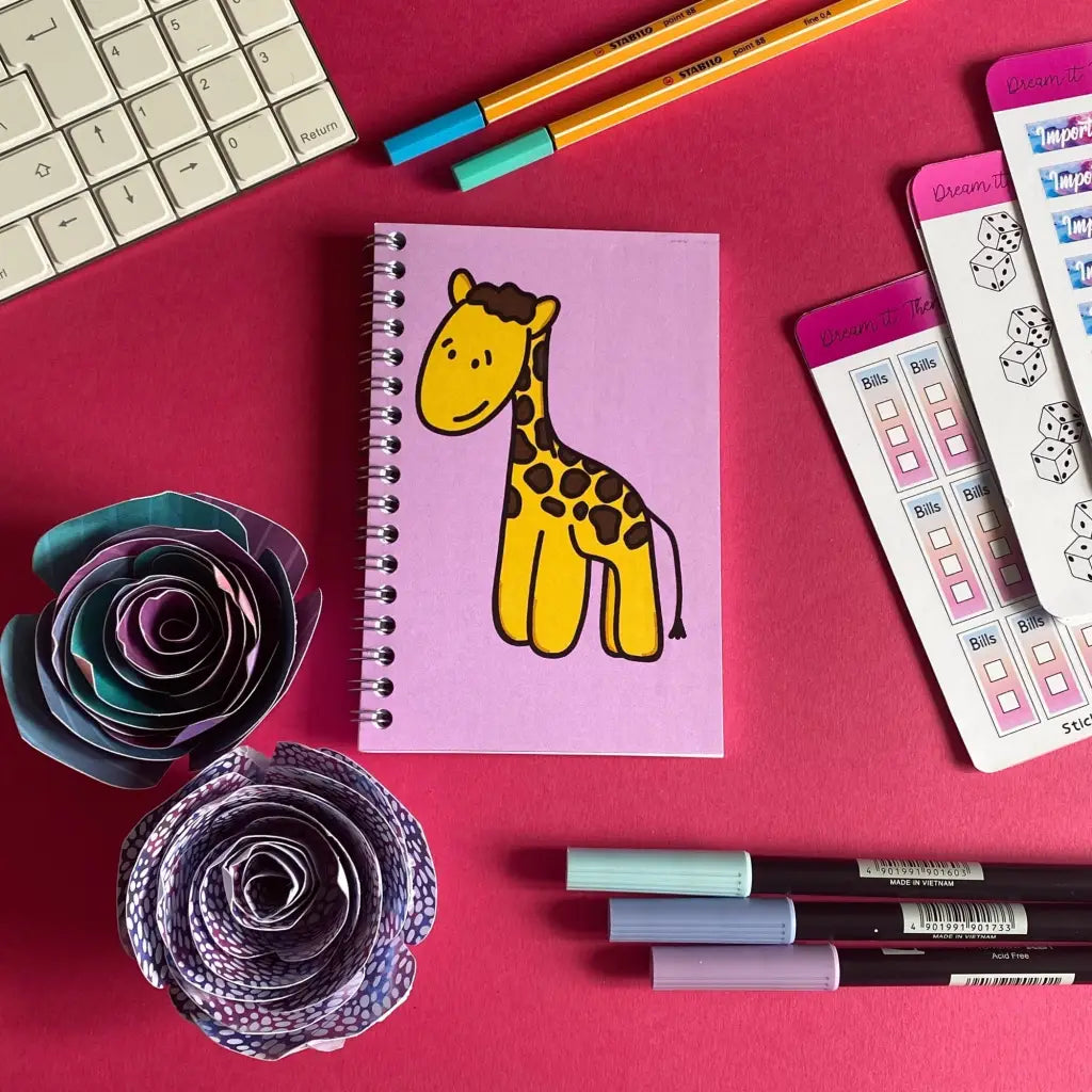 A pink desk setup featuring a Mini Happy Giraffe Notebook with a soft pink cover, surrounded by pens, markers, and paper decorations resembling flowers. A corner of a keyboard and checklist sheets can be seen. The scene is colorful and organized, with a whimsical theme.