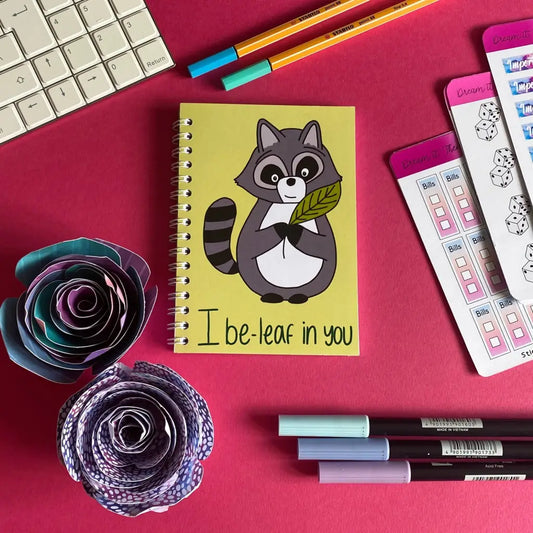 A Mini i Be-leaf in You Notebook featuring a cartoon raccoon holding a leaf with the text "I be-leaf in you" on the cover is placed on a pink surface. Surrounding the encouraging notebook are colorful pens, paper flowers, sticky notes, and the corner of a keyboard.