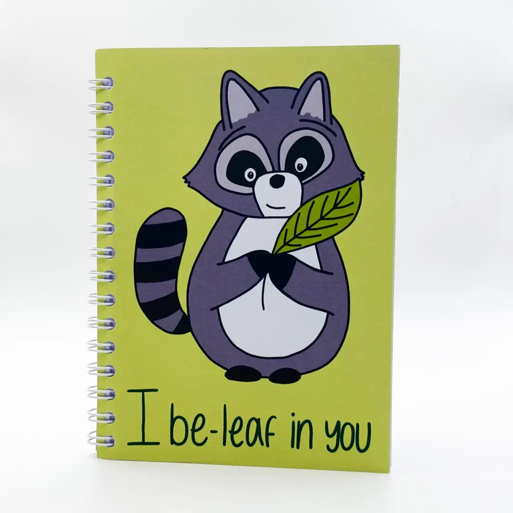 A spiral-bound Mini i Be-leaf In You Notebook with a yellow cover showcasing a cartoon raccoon holding a green leaf. The phrase "I be-leaf in you" is charmingly written on the bottom, making it a delightful raccoon notebook.