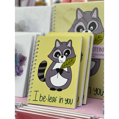 A display featuring a Mini i Be-leaf In You Notebook with a cute cartoon raccoon holding a leaf on the vibrant yellow cover. The playful pun, "I be-leaf in you," is written at the bottom. Other notebooks with different designs create a colorful background.