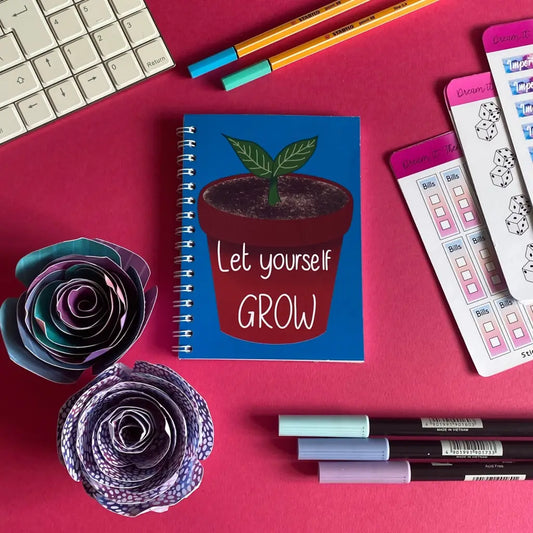 A flat lay of a desk with a Mini Let Yourself Grow Notebook featuring a sprouting plant. Surrounding items include a keyboard, colorful pens, sticky notes with checklists, and geometric paper flower crafts—all on a vibrant pink background. This setup is perfect for self-esteem boosting with its positive quote and cheerful vibes.