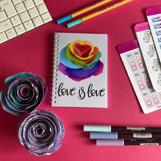 A colorful workspace featuring a Mini Love Is Love Notebook with a rainbow rose and the text "love is love" on the cover. Surrounding it are paper flowers, pens, pencils, and checklists on a pink surface next to a white keyboard, celebrating the pride community flag's vibrant spirit.