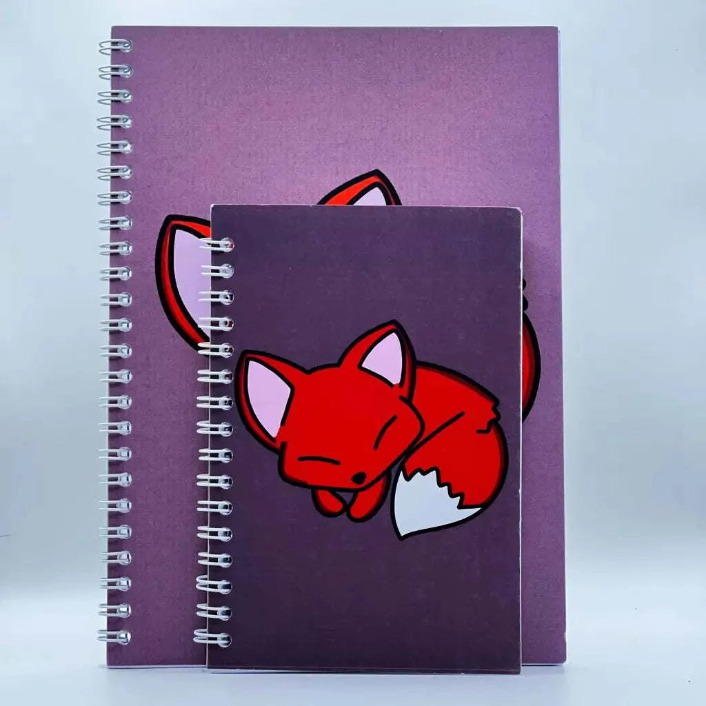 Two spiral-bound A6 plain notebooks with purple covers and an autumnal toned color palette, each featuring a cartoon image of a tired fox. The larger notebook is behind the smaller one, partially obscured, perfect for all your note-taking needs.