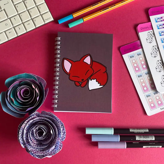 A flat lay photo on a pink background features a Mini Sleepy Fox Notebook, surrounded by colorful stationery like pens, pencils, checklists stickers, and two paper flowers. A keyboard is partially visible in the top left corner.