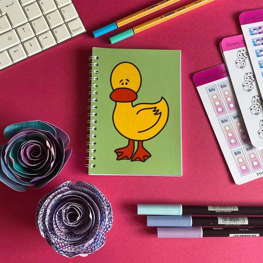 A Mini Worried Duck Notebook with a cute yellow duck on the cover is placed on a pink desk. Surrounding it are colorful pens, rolled paper flowers, a keyboard, and sheets of stickers. The 40-page scene is vibrant and organized, emphasizing creativity and productivity.