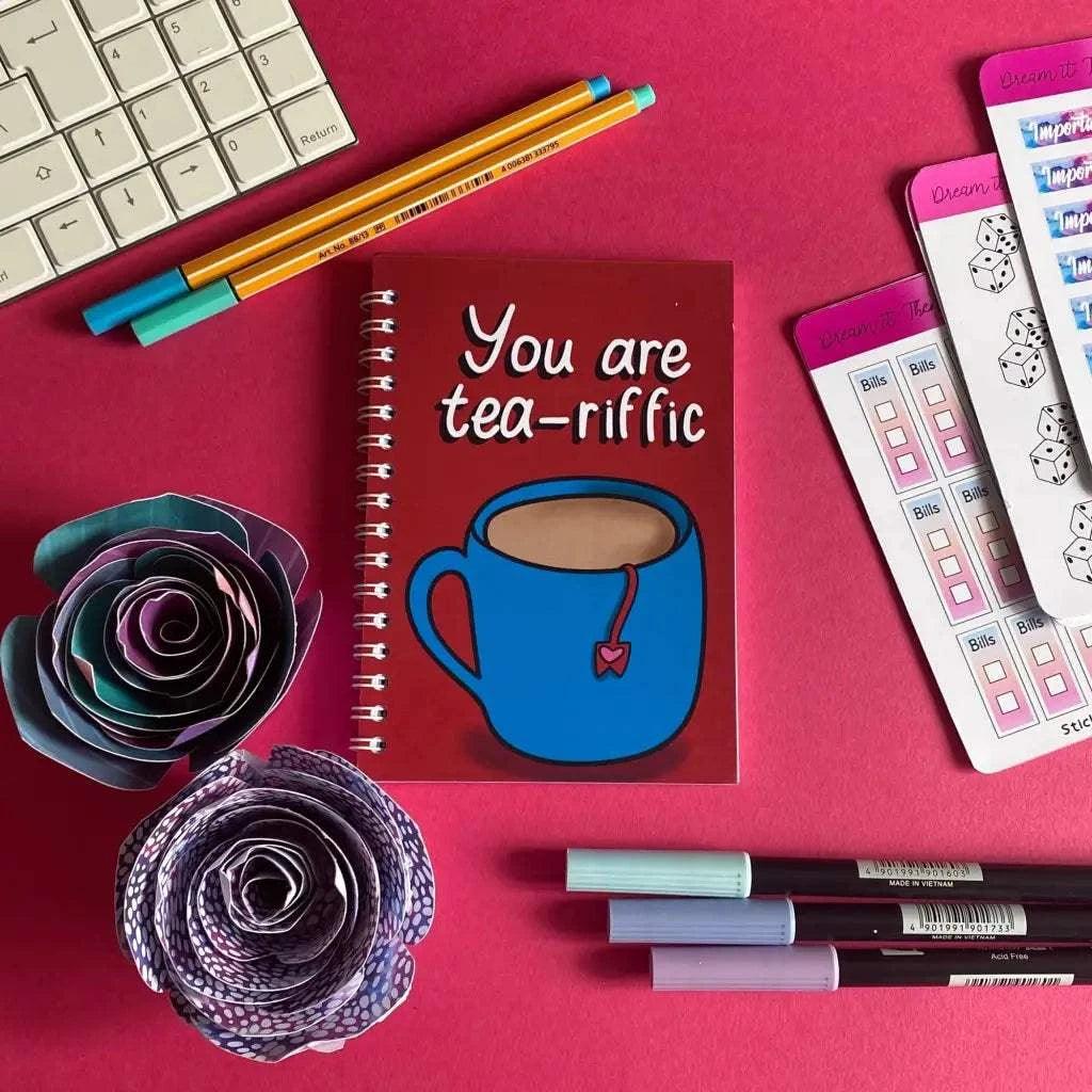 A 40-page A6 plain notebook titled "You Are Tea-riffic" on the cover, surrounded by pens, paper roses, planner stickers, and part of a keyboard on a red surface. Ideal for note-taking or planning your day.