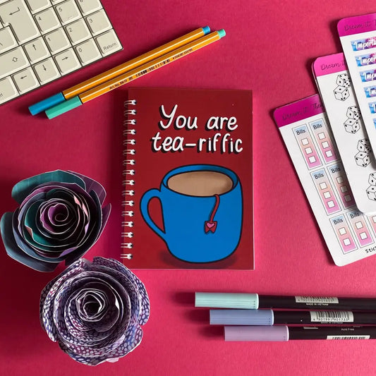 A colorful flat lay on a pink surface features a keyboard, two pencils, a Mini You Are Tea-riffic Notebook with a blue mug illustration, perfect for note-taking. A sticker sheet, three pens, and two purple paper roses complete the vibrant and neatly arranged setup.