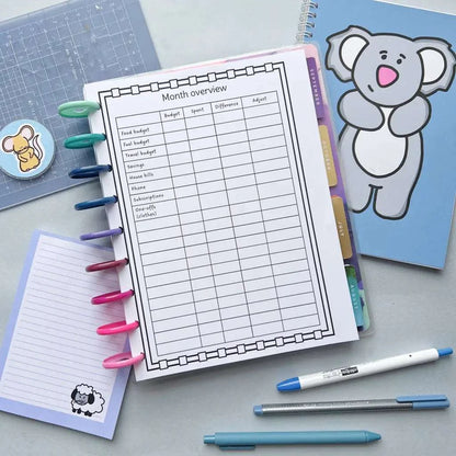 A Monthly Budget And Spending lays open on a desk, displaying a "Month overview" sheet with columns for budget, spent, difference, and adjust. Nearby are pens in various colors, a small lined notepad with a sheep illustration, a blue notebook with a koala cover, and a grid cutting mat.
