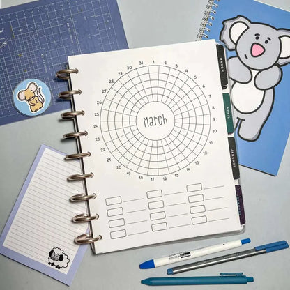 A circular March calendar template in a notebook is surrounded by various stationery items. A pen, pencil, and blue marker are below the notebook. A cute koala-themed journal for goal setting is on the right and a lined notepad with a sheep illustration perfect for a Monthly Habit Tracker is on the left.