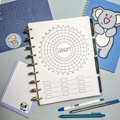 A planner open to a circular January calendar with a grid. The desk also holds a blue graph mat, a spiral notebook with a koala cover for goal setting, stickers of a koala and a lamb, a notepad with lines, and several colorful pens alongside the Monthly Habit Tracker.