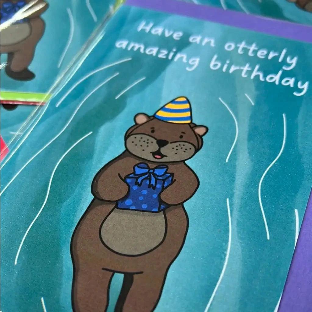 The "Otterley Amazing Birthday" card showcases a charming otter donned in a party hat and holding a gift. The cheerful illustration is set against a vibrant blue background adorned with wavy white lines, and the text reads, "Have an otterly amazing birthday." The card also features biodegradable packaging for an environmentally friendly option.