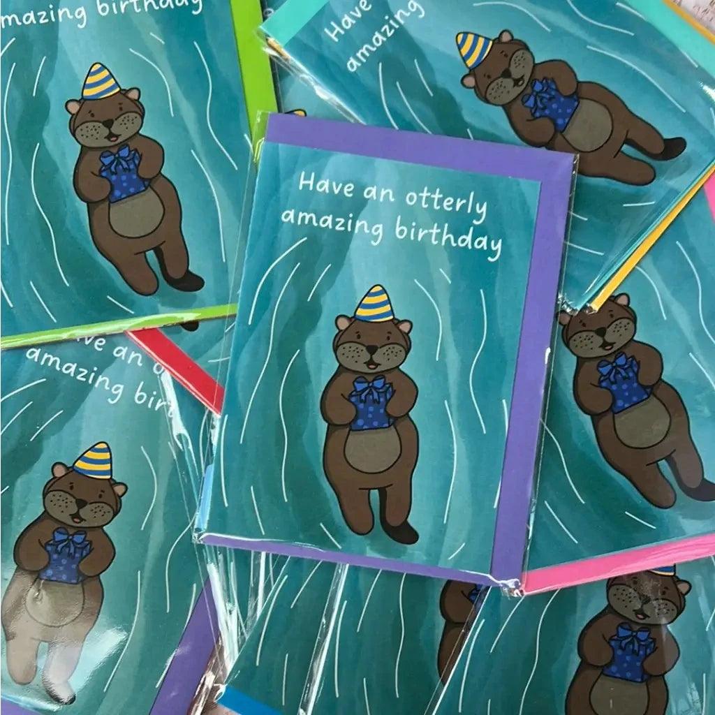 The Otterley Amazing Birthday collection consists of charming birthday cards showcasing a cute cartoon otter donned in a party hat and holding a gift. The text on the card reads, "Have an otterly amazing birthday." Available with green, blue, pink, and purple borders, these cards are also packaged in biodegradable material.