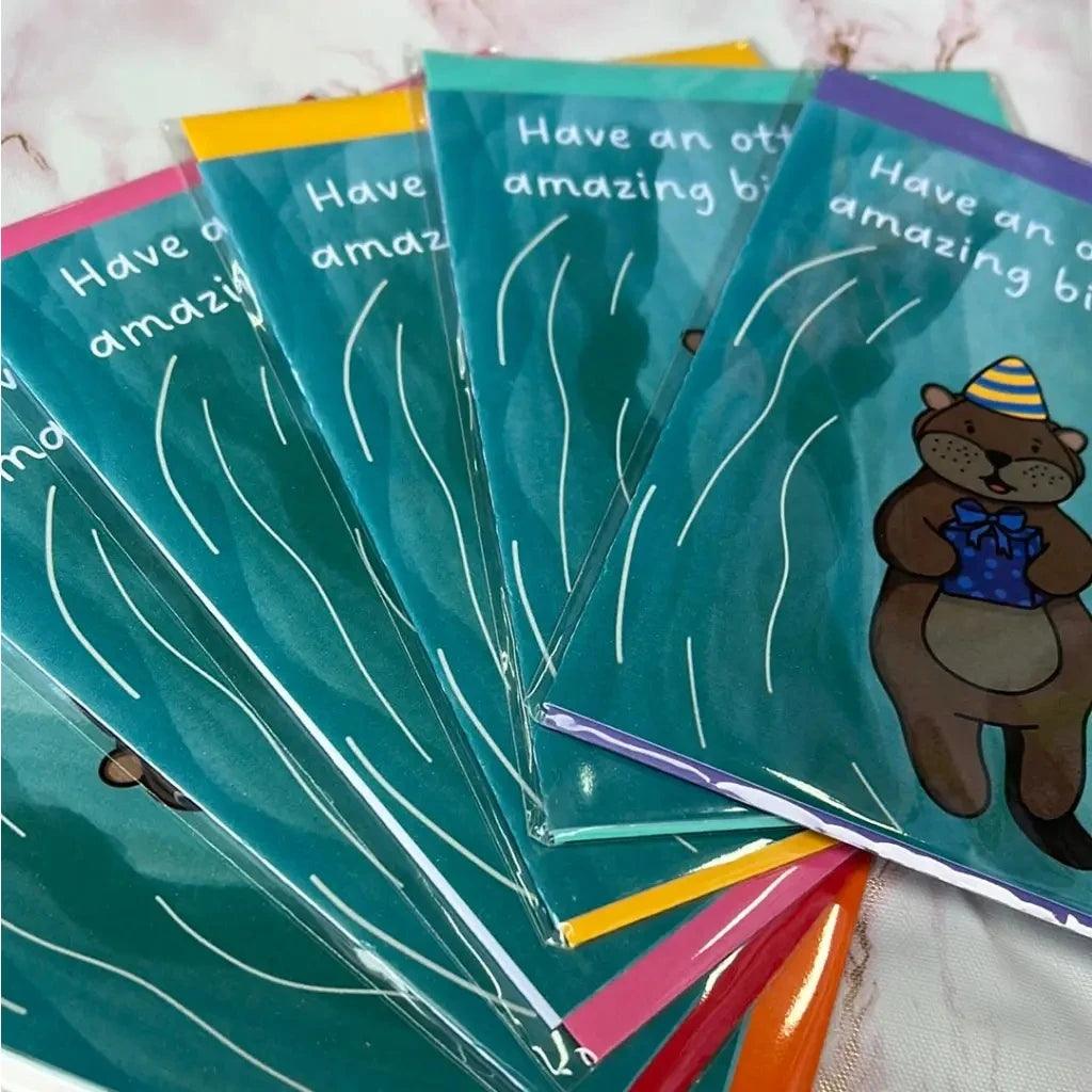 Seven colorful "Otterley Amazing Birthday" cards are displayed in a fanned-out arrangement. Each happy birthday card features an adorable otter holding a wrapped gift and wearing a party hat. The text on the cards reads, "Have an otterly amazing birthday!" They come in biodegradable packaging for an eco-friendly touch.