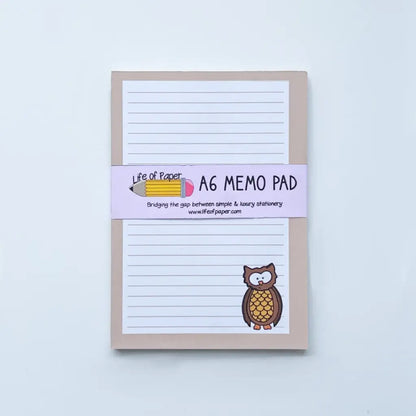A beige A6-sized memo pad with lined paper is shown against a white background. The cover features a small owl illustration at the bottom right and a pink band around the pad with text and a pencil illustration, labeled "Owl Memo Pad." This mini memo pad is fountain pen friendly.