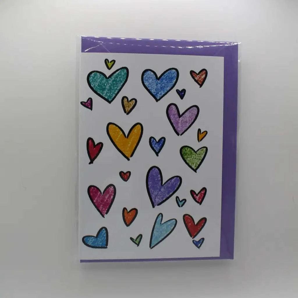 The Rainbow Hearts Greeting Card is a biodegradable greeting card with a purple border, adorned with multiple colorful hand-drawn hearts of various sizes on the front. It's perfect for sending any note.