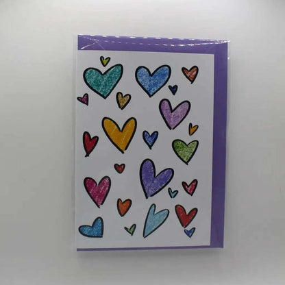 The Rainbow Hearts Greeting Card is a biodegradable greeting card with a purple border, adorned with multiple colorful hand-drawn hearts of various sizes on the front. It's perfect for sending any note.