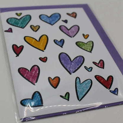 The Rainbow Hearts Greeting Card is a biodegradable, white card featuring an array of colorful, hand-drawn hearts in various sizes and shades, set against a purple background.