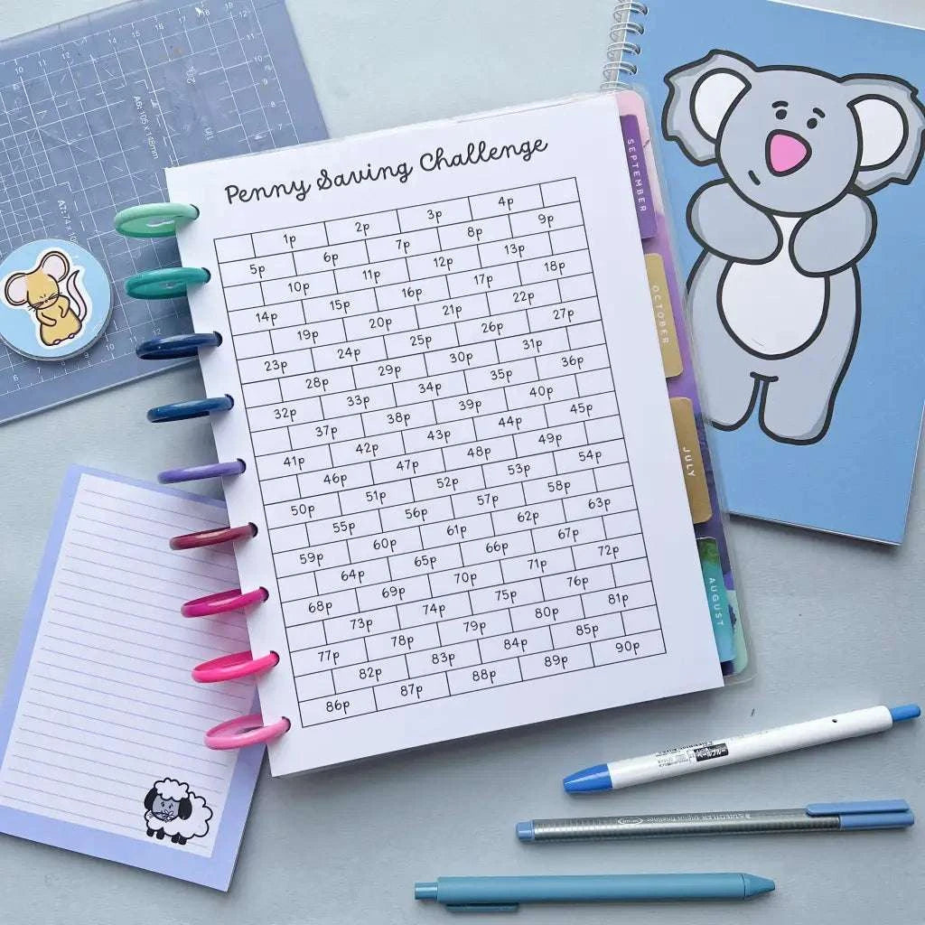 A Penny Saving Challenge in a planner with colored tabs is on a table, surrounded by an array of stationery including blue-handled pens, colored markers clipped on the left, a notepad with a sheep illustration, and a binder with a koala cover. Perfect for visual savings tracking on your planner pages.