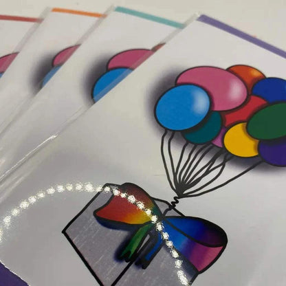 Inclusive Rainbow Parcel Greeting Cards featuring an illustration of a gift box tied with a rainbow ribbon and colorful balloons rising from it. Four biodegradable Rainbow Parcel Greeting Cards are fanned out, showcasing the vibrant design on a white background, with some glare visible.