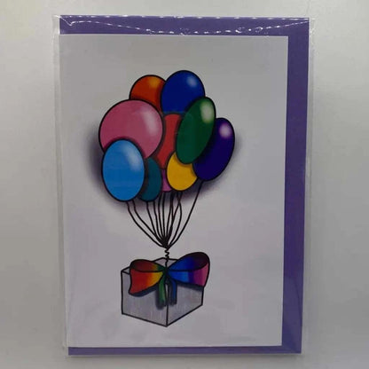 The Rainbow Parcel Greeting Card showcases an illustration of a transparent gift box adorned with a colorful ribbon. Attached to the gift box are ten vibrant balloons in various shades, such as pink, blue, red, yellow, green, and purple. The inclusive design is completed with a biodegradable purple envelope.