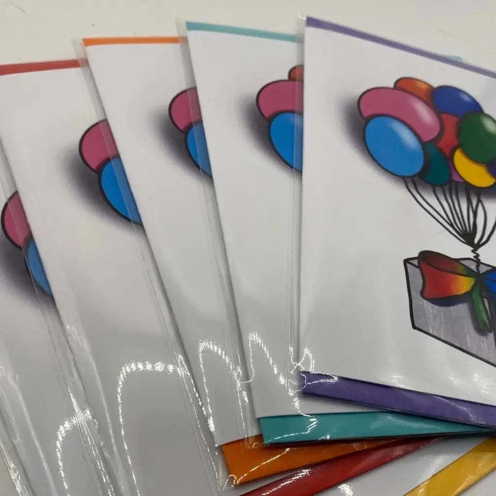 A set of Rainbow Parcel Greeting Cards, featuring a stunning illustration of balloons tied to a gift box, are biodegradable and partially fanned out to showcase their vibrant borders in various colors including red, orange, yellow, and purple.