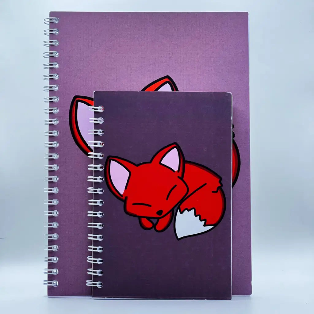 Two spiral-bound notebooks are stacked upright against a light background. The larger notebook has a purple cover, and the smaller, autumnal-toned Sleepy Fox Notebook in front features a cute red fox cartoon illustration. Both notebooks, with 40 pages each, have similar spiral binding on the left side.