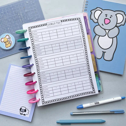 A budgeting planner lies open on a desk with the heading "Snowball Monthly Payment Log." Multicolored pens are clipped to the left margin. Surrounding the planner are a notepad with a sheep illustration, a blue notebook with a koala illustration, and various stationery items.