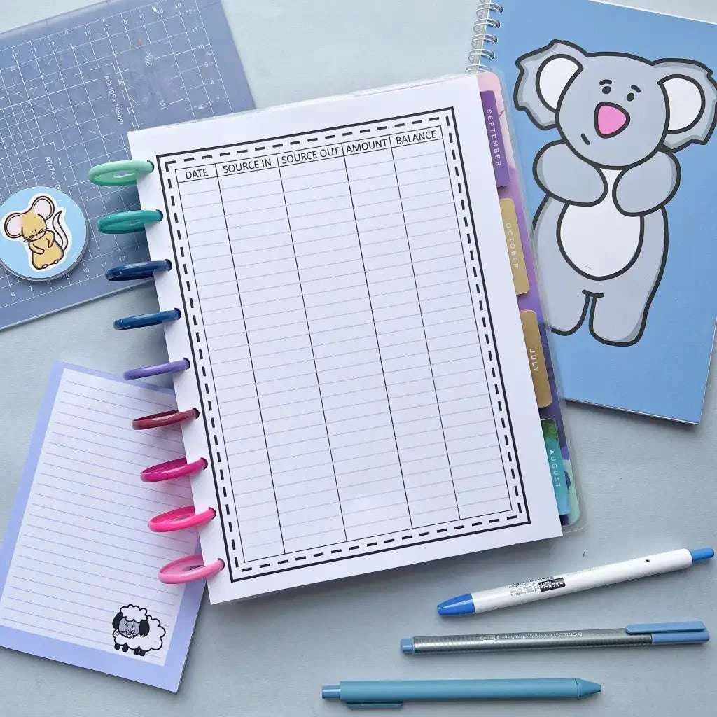An image of a workspace featuring a balance ledger sheet on top of a ring binder, surrounded by colorful pens. Amidst the array, there's a Spending Tracker Page with a cartoon koala cover and a small notepad with a sheep illustration, perfect for monthly budgeting and tracking spending habits with assorted stationery.