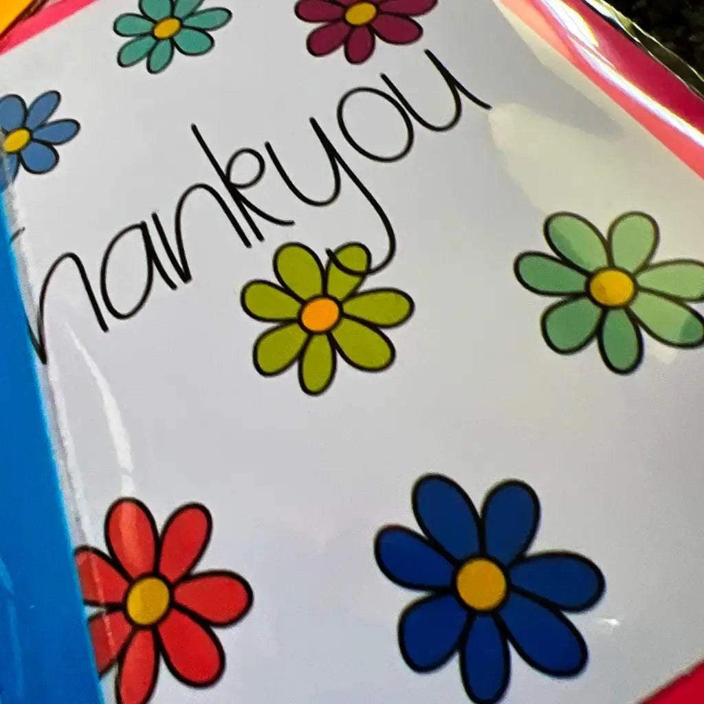 The Thank You Card features the words "thank you" in a playful font, surrounded by bright, illustrated flowers in red, blue, yellow, green, and more. For eco-friendly gifting, it comes packaged in a biodegradable cellophane bag. The edges are partially visible with a blue stripe on the left side.