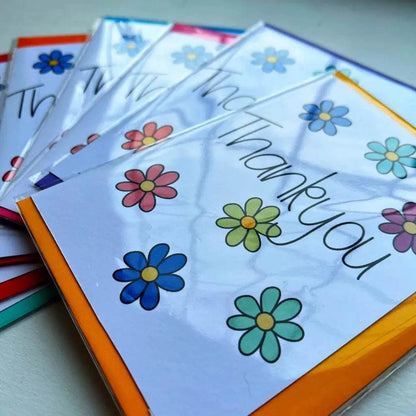 A close-up shot showcases several vibrant "Thank You Card" designs featuring flower motifs, arranged fan-like. Each card displays the words "Thank You" in an elegant script amidst colorful flowers. The eco-conscious presentation is enhanced by some cards being enclosed in biodegradable cellophane bags.