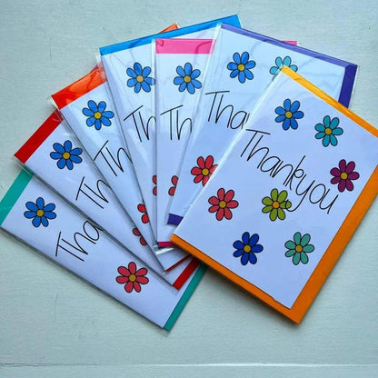 The Thank You Card set includes six vibrant and beautifully designed cards, each showcased in a fanned-out arrangement. Every card displays the words "Thank you" elegantly written in cursive, surrounded by a cheerful design of brightly colored flowers. These cards feature borders in various colors such as orange, purple, pink, blue, red, and green. They come packaged in an environmentally friendly biodegradable cellophane bag.