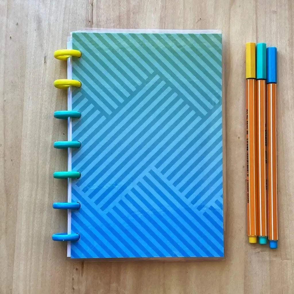 A Tiny Notes Notebook with a blue geometric patterned cover sits on a wooden surface. The notebook has colorful rings along its spine. To the right of the notebook, there are four orange and blue pens neatly lined up, making it MAMBI Happy Planner compatible and fountain pen friendly.
