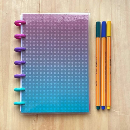 A colorful, fountain pen friendly notebook with a gradient cover from pink to blue, featuring white polka dots, lies on a light wooden surface. To the right of the Tiny Notes Notebook, there are three markers in pink, yellow, and blue, aligned neatly.